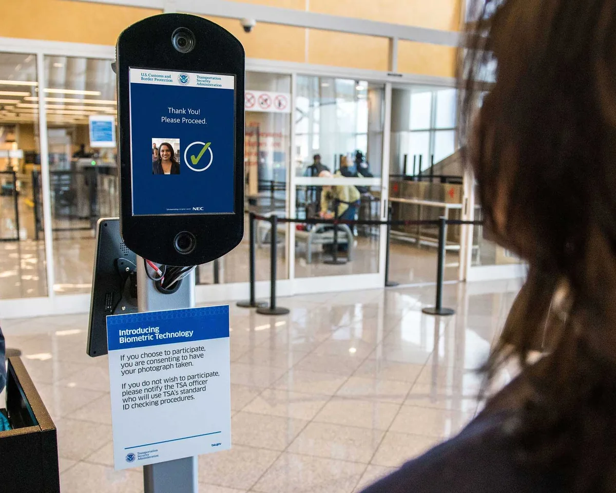 An airport face-scan kiosk showing a traveler&rsquo;s photo, a green check mark and &ldquo;Thank you! Please proceed,&rdquo; above a sign explaining that taking part means consenting to be photographed.